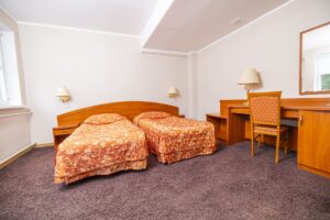 dwa łóżka z pomarańczową narzutą, biurko z krzesłem po prawej stronie