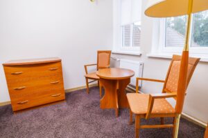 komoda, stolik z dwoma pomarańczowymi krzesłami, lampka stojąca