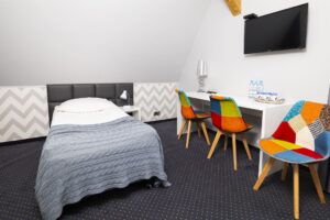 pokój z łóżkiem pojedynczym, białe biurko, kolorowe krzesła i telewizor